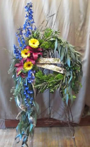 Wreath Tribute with Delphinium, Asiatic Lilies and Large Gerbera Daisy flowering on a wreath of mixed greens and field grasses with draping Eucalyptus